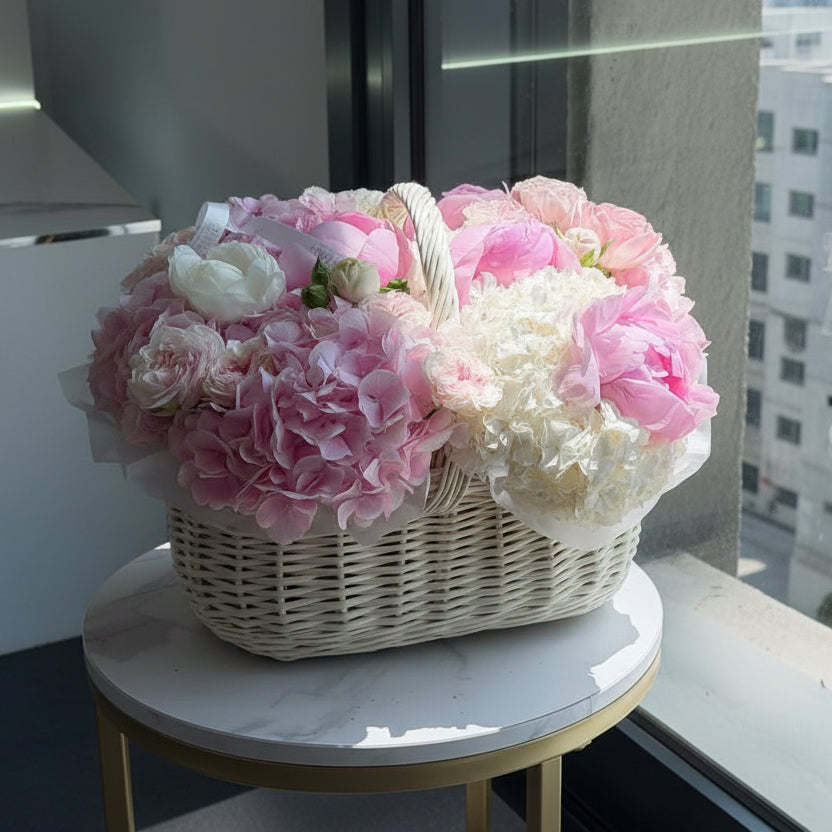 A wicker basket filled with pink and white peonies and hydrangeas, displayed on a round marble-top side table.